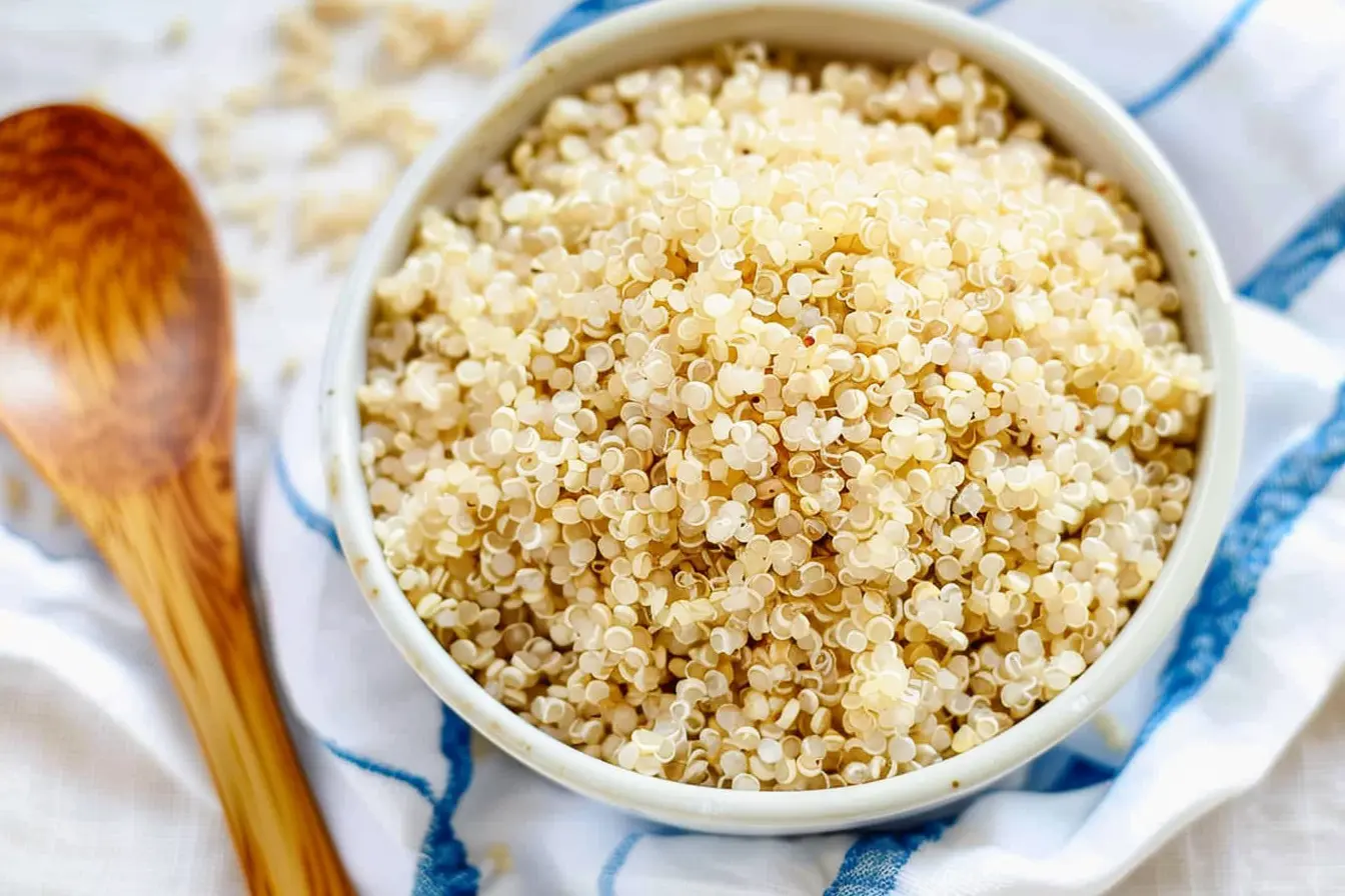 A bowl of fluffy, cooked quinoa with a wooden spoon on the side, displayed on a white and blue kitchen towel, showcasing a nutritious alternative to rice