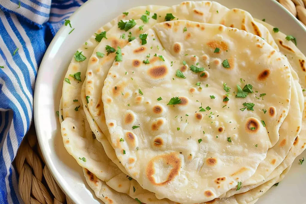 A plate of freshly made unleavened bread, lightly charred with golden brown spots and garnished with chopped cilantro. The flatbread is soft and thin, served on a white plate with a blue-striped napkin beside it.