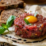 Beef tartare topped with a raw egg yolk and garnished with fresh basil leaves, served on parchment paper with slices of seeded bread in the background and a drizzle of balsamic reduction.