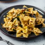 Plate of farfalle pasta tossed with sautéed zucchini, mushrooms, and red pepper flakes, served on a dark plate with a fork on the side and a green cloth in the background.