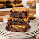 Stack of rich, chocolatey brookies with layers of brownie and chocolate chip cookie, arranged on a white plate with chocolate chips scattered around and a glass of milk in the background.